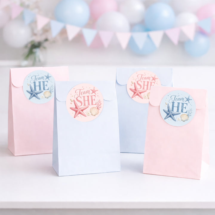 Four pastel party favor bags standing on a white surface, decorated with round stickers that read 'Team HE' and 'Team SHE' with starfish and shell motifs. The bags are pink, light blue, and pale blue, with a soft pink and blue balloon banner blurred in the background.