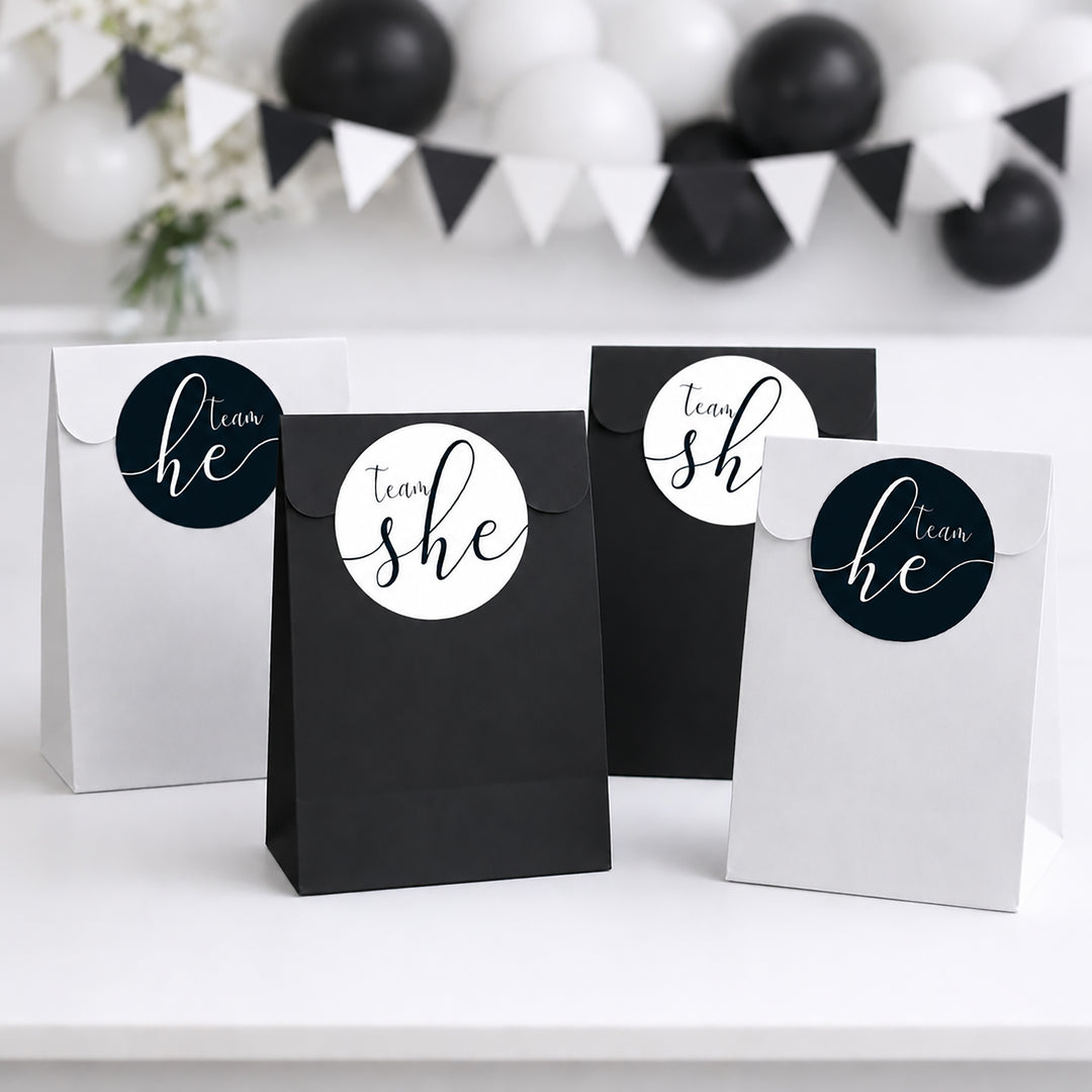 Four paper party favor bags in black and white, each with a circular custom name sticker reading 'team she' or 'team he' in white script on a dark blue background, arranged on a white surface with blurred black and white balloons in the background.