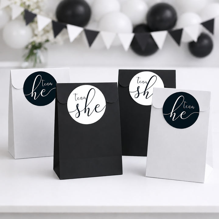 Four paper party favor bags in black and white, each with a circular custom name sticker reading 'team she' or 'team he' in white script on a dark blue background, arranged on a white surface with blurred black and white balloons in the background.