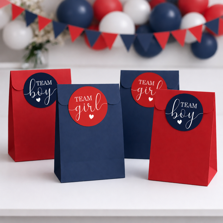 Four paper party favor bags in red and navy blue stand on a white surface, each with a round sticker reading TEAM boy or TEAM girl with a small heart. Red, white, and blue balloons and bunting are blurred in the background.